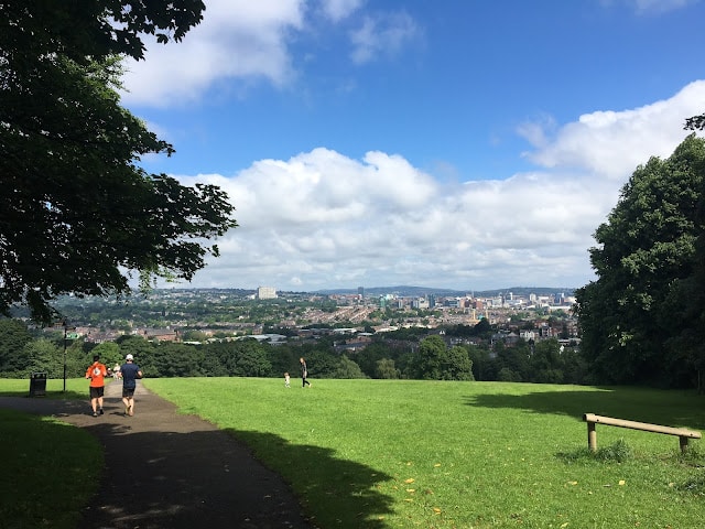 Two people jog on a paved path through a green park with trees in Yorkshire, overlooking a cityscape under a partly cloudy blue sky. A few more people are visible in the distance, enjoying one of the great places to raise children.