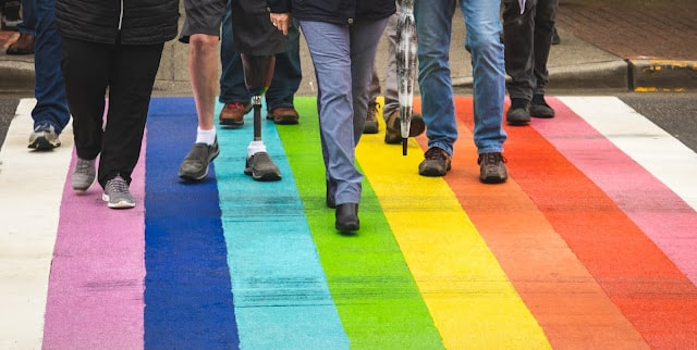 A group of people walking across a rainbow-coloured pedestrian crossing, symbolising LGBTQ+ pride and diversity—an everyday scene that helps teach children inclusivity through visible support in our communities. Only the lower half of their bodies and legs are visible.
