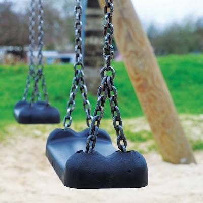 Close-up of two empty black swings hanging by metal chains at a playground, with a sandy ground and grassy area in the background. This scene highlights socially distanced playtime, with the focus on the nearest swing.