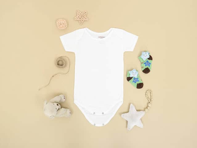 A plain white baby vest is laid flat on a beige surface, surrounded by baby socks, soft toys, and cute decor—perfect for welcoming a newborn or inspiring baby celebration ideas.