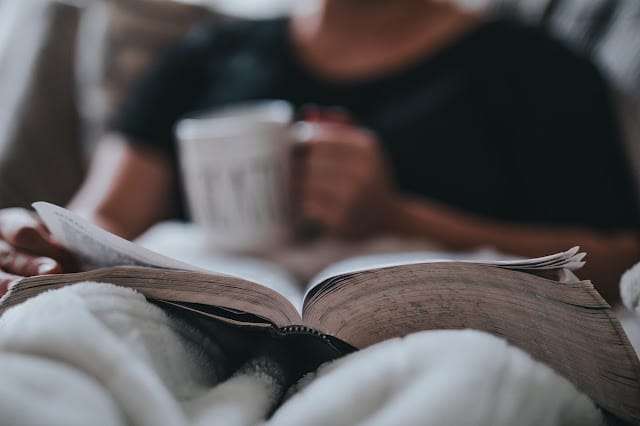 A person enjoying some self-care sits cosy under a blanket, holding a mug and reading an open book on their lap. The focus is on the book, capturing a peaceful moment of personal time with the reader softly blurred in the background.
