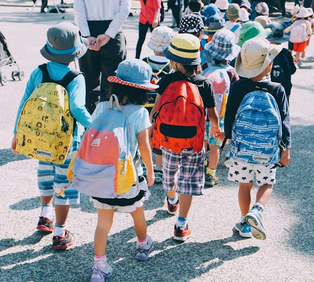A group of young children dressed for going back to school, wearing colourful hats and rucksacks, walk together outdoors on a sunny day, with an adult visible ahead of them.