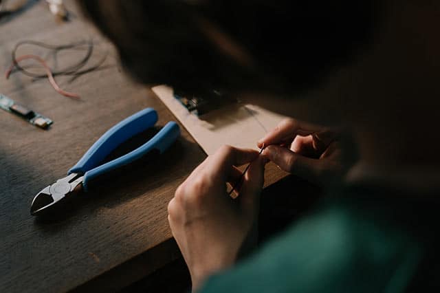 A person working with small electronic components at a wooden table, holding wires in their hands, with blue-handled wire cutters and other essential tools from their tool box scattered nearby.
