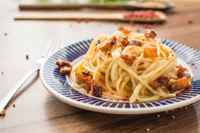 A plate of spaghetti topped with crispy bacon pieces and a light dusting of seasoning, showcasing simple kitchen skills, is placed on a wooden table with a fork beside the plate.