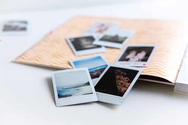 Several small instant photos are spread out on a white surface next to a peach-coloured patterned photo album, with some images partially tucked inside—showing a simple way for how to make memories last.