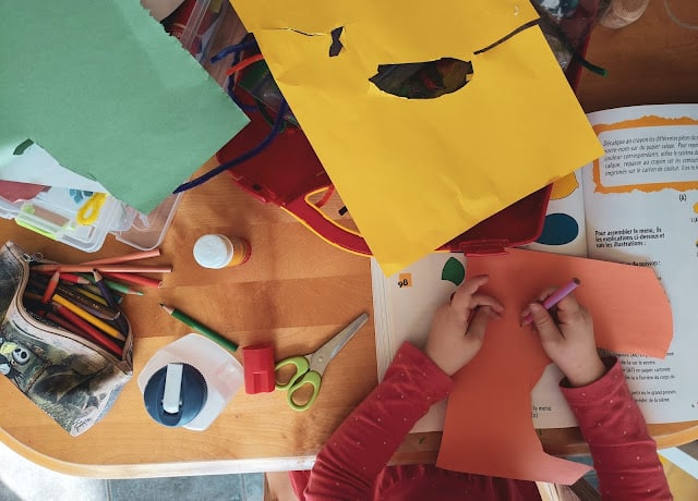 A child’s hands cut orange paper with scissors on a cluttered table, surrounded by coloured paper, crayons, glue, and an open book—a perfect scene for entertaining children with creative crafting ideas on a rainy day.