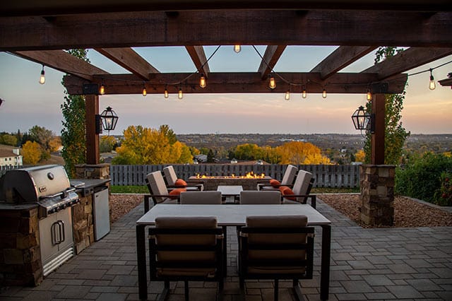 A cosy patio with a modern pergola, fairy lights, a barbecue, a dining table with chairs, and a seating area around a firepit overlooks a scenic landscape with autumn trees and a distant town at sunset.