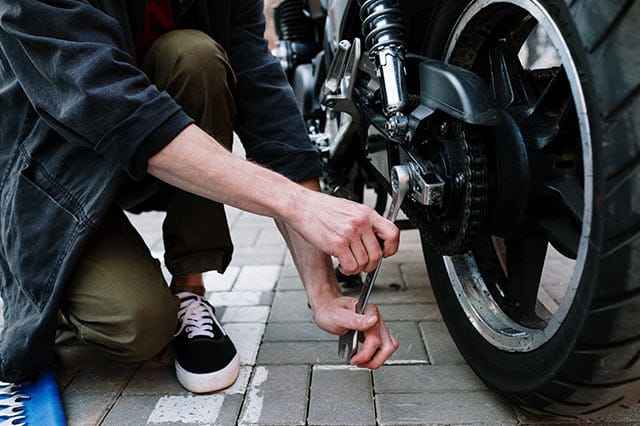 A person kneels on the ground using a spanner and essential tools to repair or adjust the rear wheel of a motorbike parked on a paved surface.