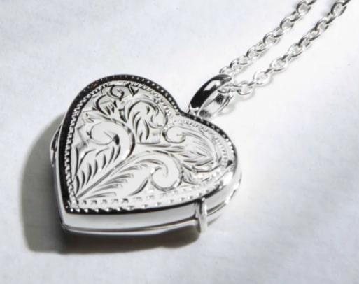 A close-up photo of a silver heart-shaped locket with an ornate, engraved floral design on the front, attached to a delicate chain—an elegant choice among Christmas gifts for adults—resting on a white surface.