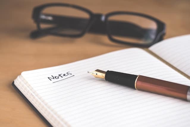 A pair of glasses sat next an open book that says Notes on the top of the page with a black pen sat on top of the book