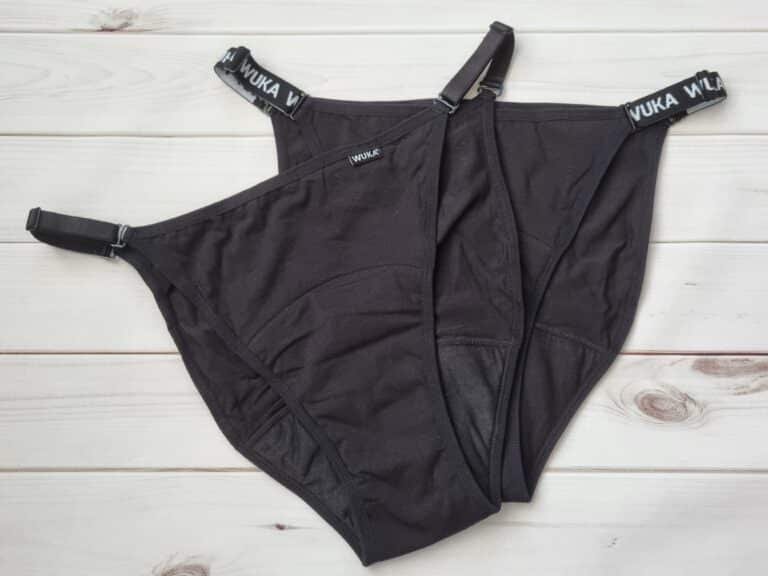 A pair of black Wuka Flex period knickers with adjustable straps and "WUKA" branding on the waistband, laid flat on a white wooden surface.