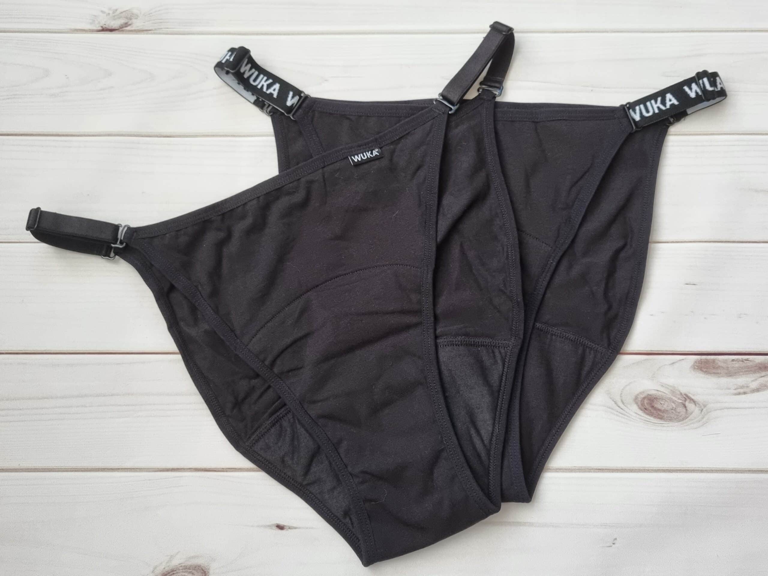 A pair of black Wuka Flex period knickers with adjustable straps and "WUKA" branding on the waistband, laid flat on a white wooden surface.