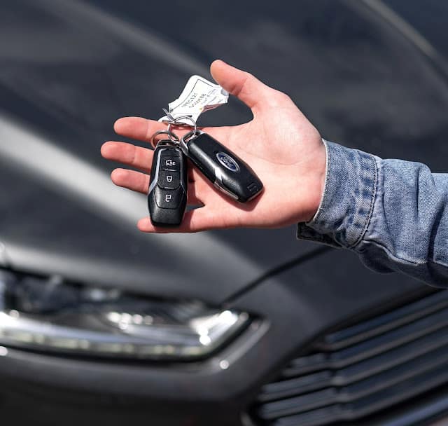 A person wearing a denim jacket holds two car key fobs and a key tag in their hand, with the front of a dark-coloured car visible in the background—perfect for illustrating things to consider when buying a new car.
