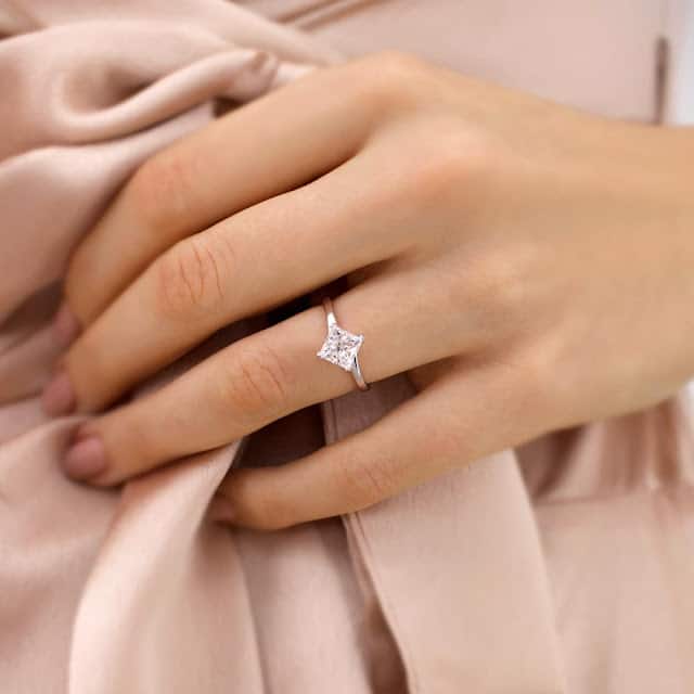 A hand wearing a silver ring with a square-cut diamond rests gently on soft, light pink fabric, suggesting elegance and sophistication—an exquisite choice inspired by a guide to lab-grown diamonds.