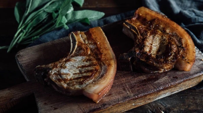 Two cooked pork chops with a golden-brown crust rest on a wooden chopping board, making this meal one of the best last-minute Father’s Day gift ideas for enjoying together. Fresh green herbs and a dark cloth set the perfect backdrop.