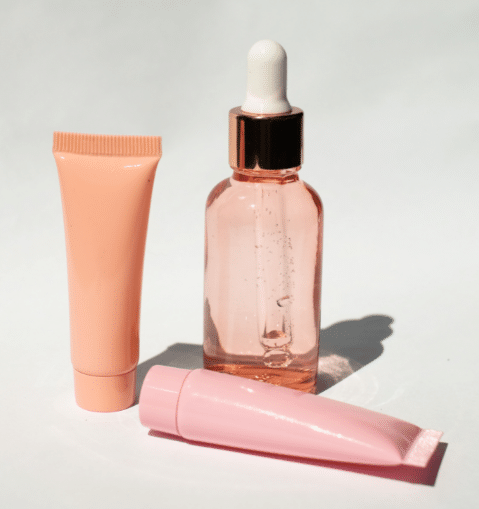 Three skincare products are displayed: a pink squeeze tube, a clear glass bottle with a dropper cap containing a liquid, and a light pink squeeze tube lying down—like freebies you might discover with tips and tricks for finding freebies on the Internet.