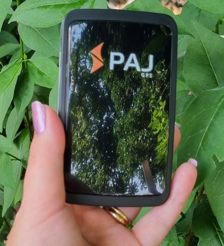 A hand holding a black PAJ GPS device outdoors during a family holiday, with green leaves in the background and the reflection of trees visible on the device’s screen.