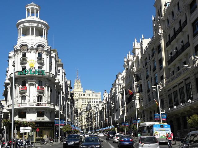 A wide street in Madrid lined with ornate, historic buildings is busy with cars and pedestrians. The sky is clear and blue. A prominent white building with a "Rolex" sign stands out on the left.
