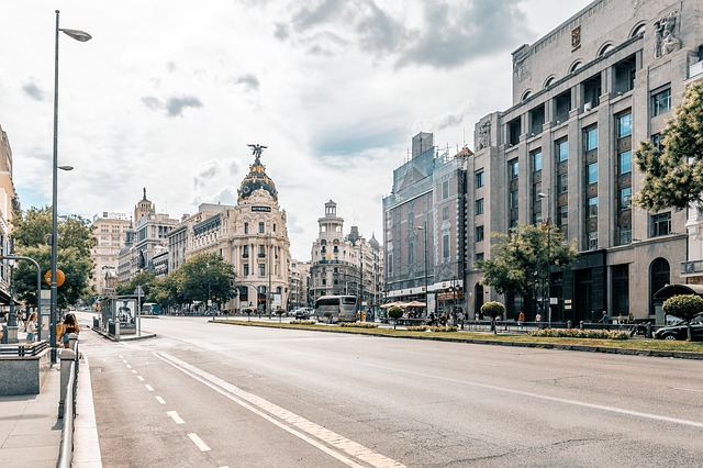 A wide city street in Madrid, Spain, features historic buildings like the ornate Metropolis Building. Trees, lamp-posts, and a few people are visible under a cloudy sky in this vibrant Madrid scene.