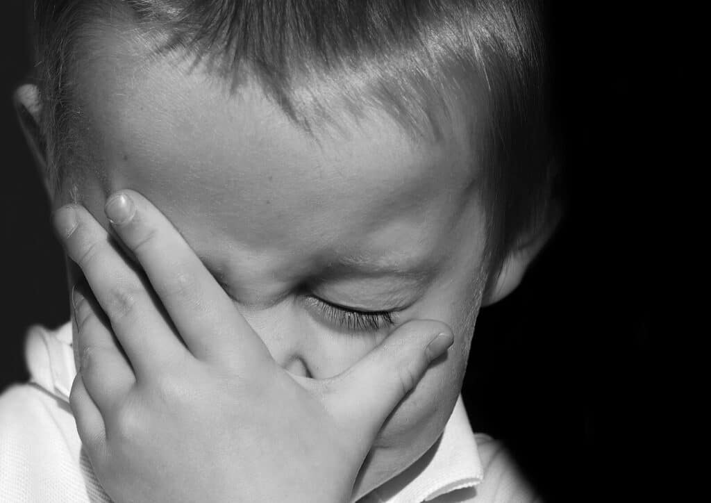 A young child covers their face with one hand, eyes closed tightly, expressing distress or frustration. The image is in black and white, highlighting the child's emotion.