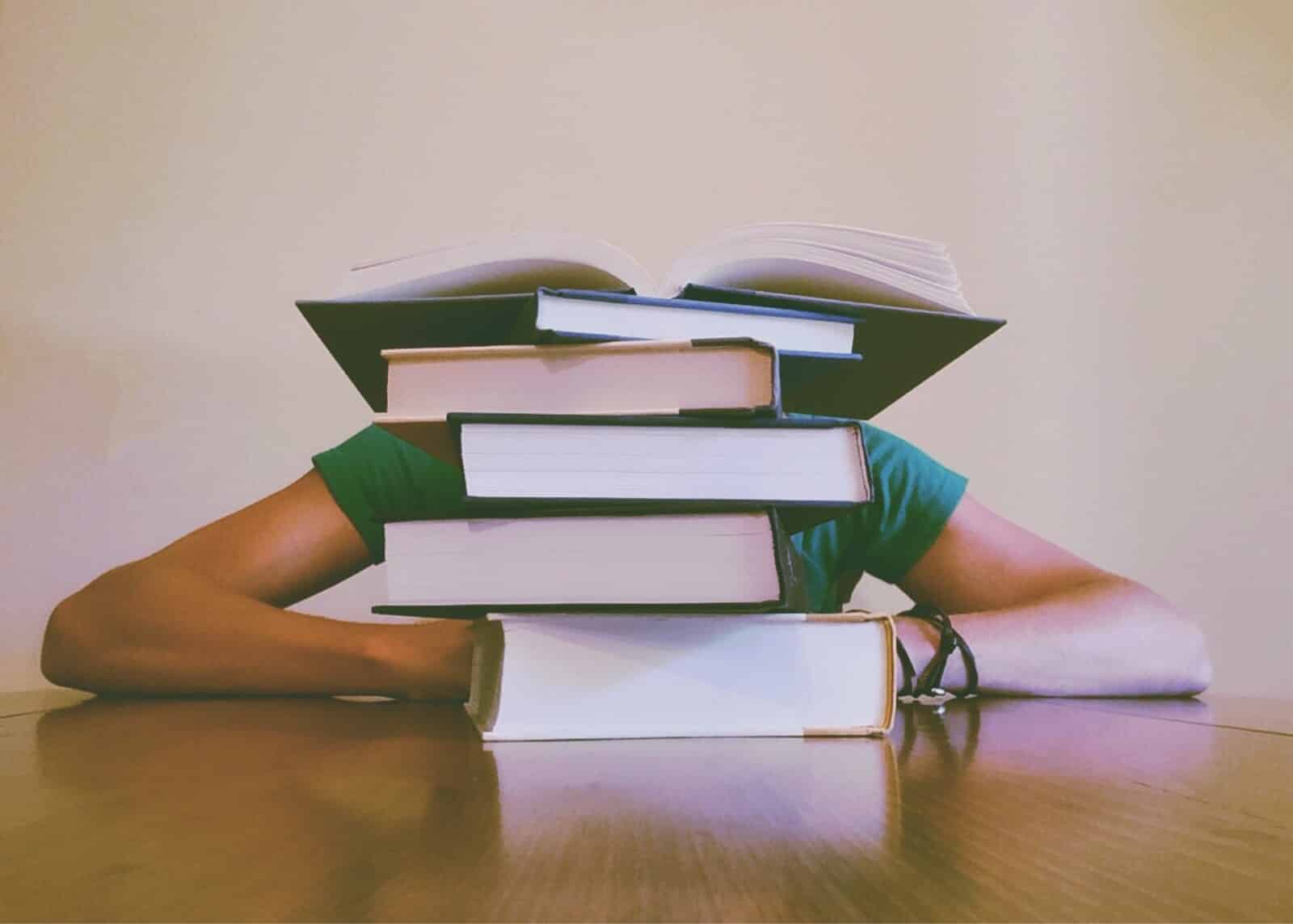 A person wearing a green shirt sits at a table, mostly hidden behind a tall stack of books with one open book on top, suggesting intense studying or reading.
