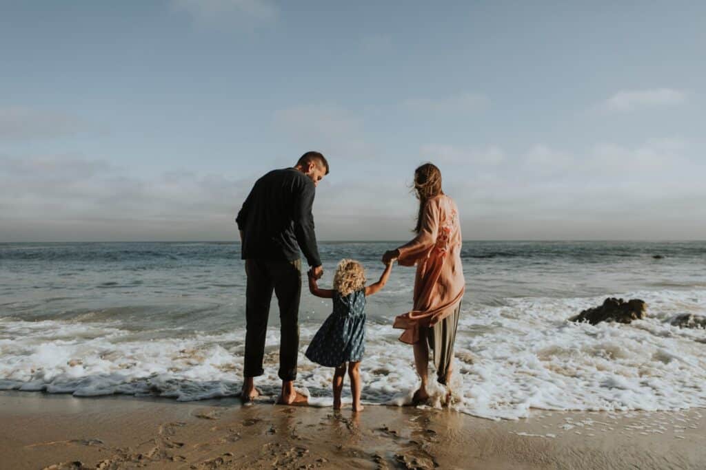 A family of three—two adults and a young child—stand barefoot at the edge of the sea, holding hands and looking out at the water, with gentle waves approaching their feet on a sandy beach.