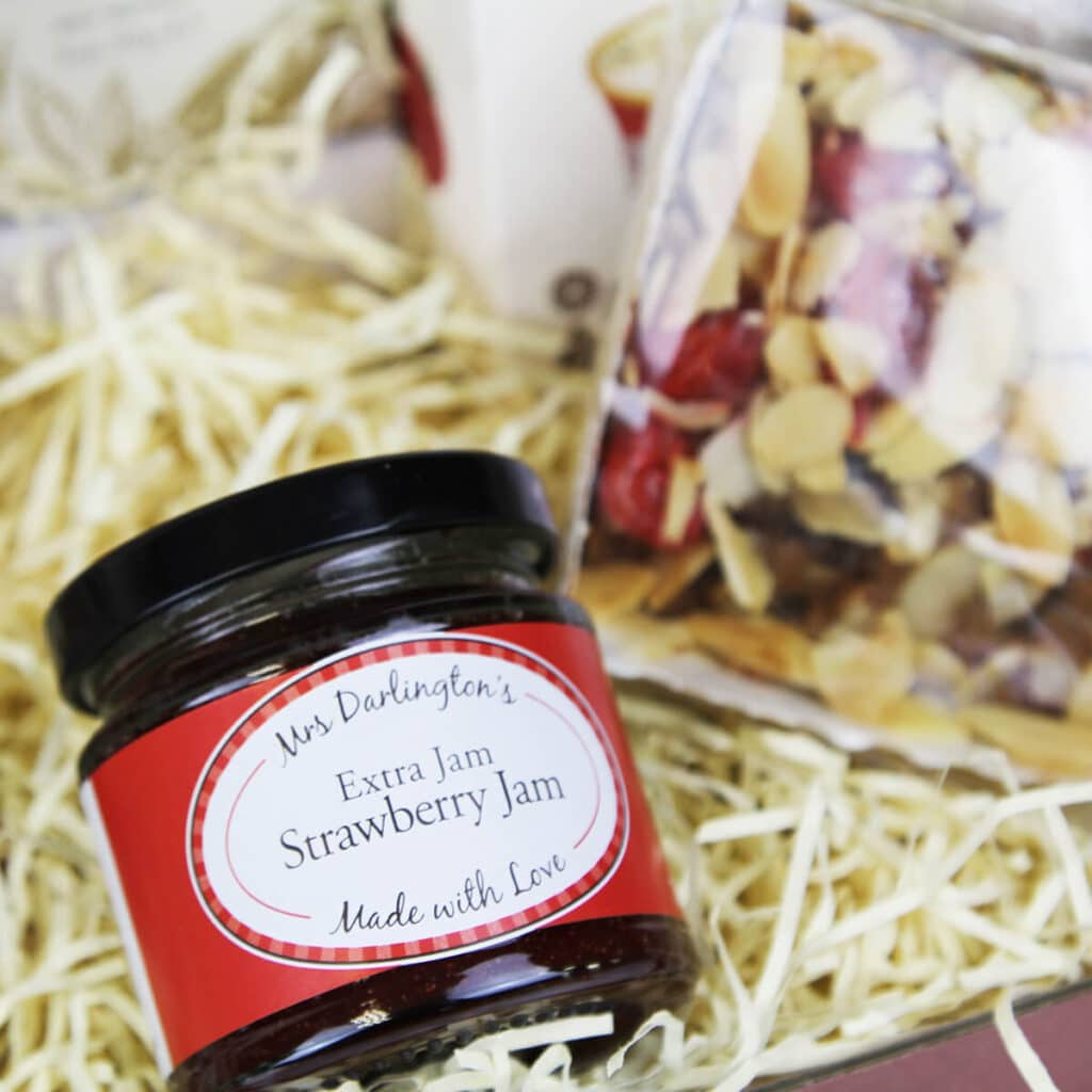 A jar of Mrs Darlington's Extra Jam Strawberry Jam with a red label sits on straw packaging next to a clear packet of almond-topped pastries, perfect for an afternoon tea hamper.