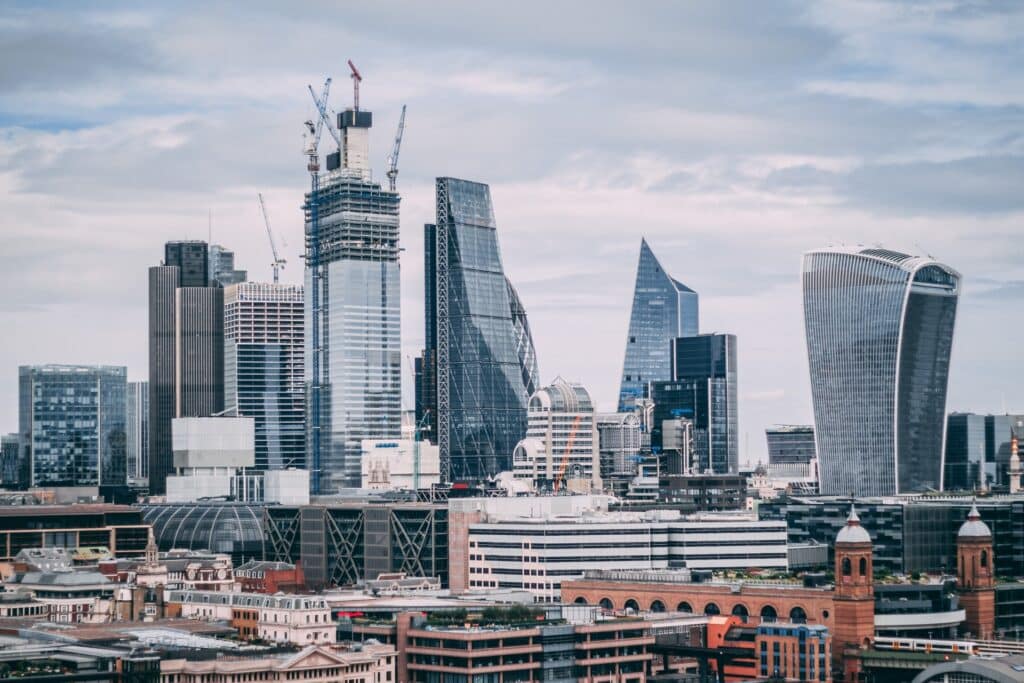 London city skyline with modern skyscrapers, cranes on buildings under construction, and a mix of glass and steel architecture beneath a cloudy sky. Older brick buildings line the foreground—an iconic view for those seeking survival tips to enjoy your first holiday in London.