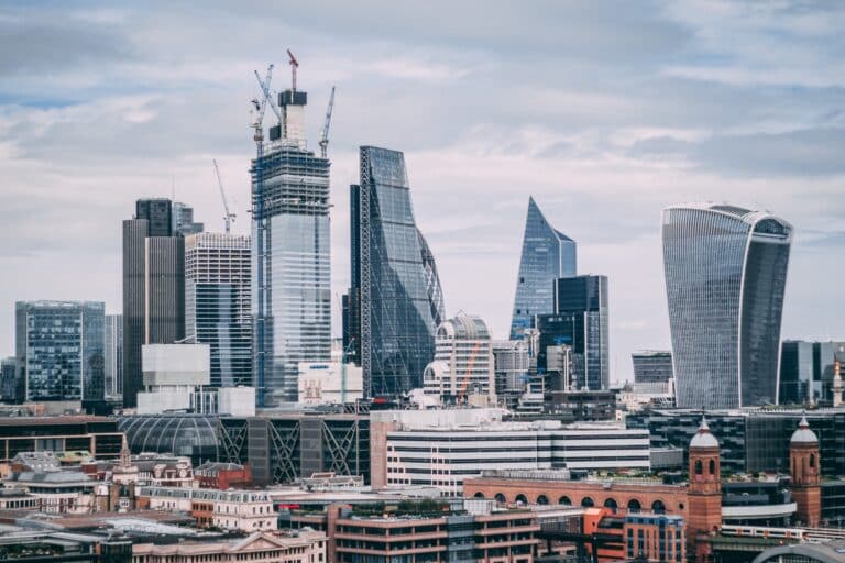 London city skyline with modern skyscrapers, cranes on buildings under construction, and a mix of glass and steel architecture beneath a cloudy sky. Older brick buildings line the foreground—an iconic view for those seeking survival tips to enjoy your first holiday in London.