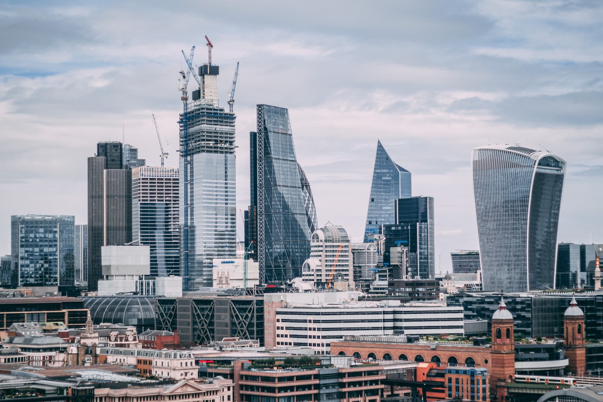 London city skyline with modern skyscrapers, cranes on buildings under construction, and a mix of glass and steel architecture beneath a cloudy sky. Older brick buildings line the foreground—an iconic view for those seeking survival tips to enjoy your first holiday in London.