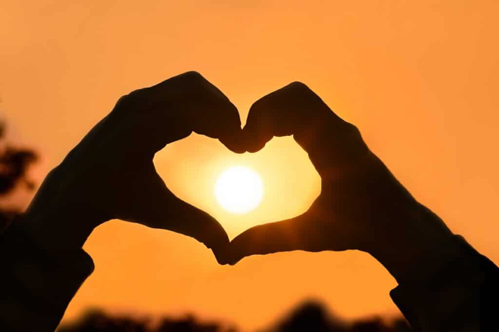 Two hands form the shape of a heart with their fingers, framing the setting sun against an orange sky—showcasing how your clothing can complement life’s most beautiful moments.