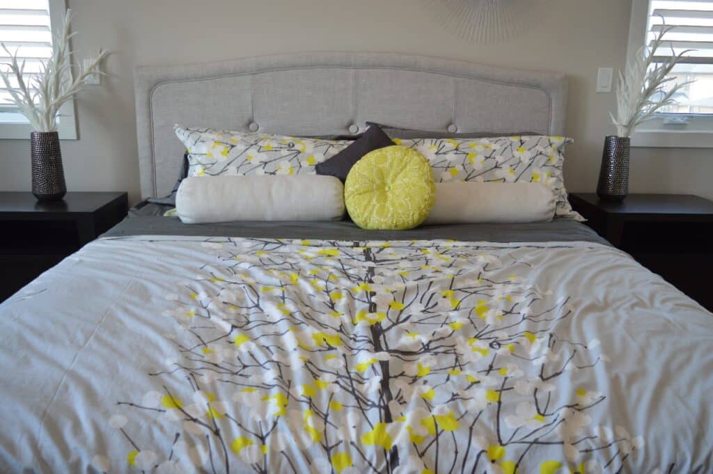 A neatly made bed with a light grey upholstered headboard, floral-patterned bedding in yellow, white, and grey—showcasing one of five ways to create extra space in your bedroom—plus decorative cushions and matching bedside tables topped with vases of white branches.