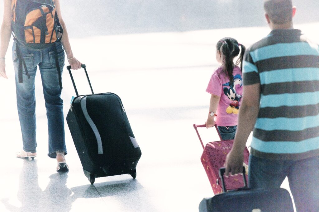 Three people, including a child with a Minnie Mouse shirt and a small pink suitcase, walk through a bright airport terminal on their family holiday, each pulling wheeled luggage.