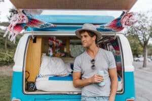 A man in a grey hat and t-shirt leans against the open back of a blue van, holding a can. The van is decorated with blankets and pillows, creating a cosy camping vibe. Trees and a car park are in the background.