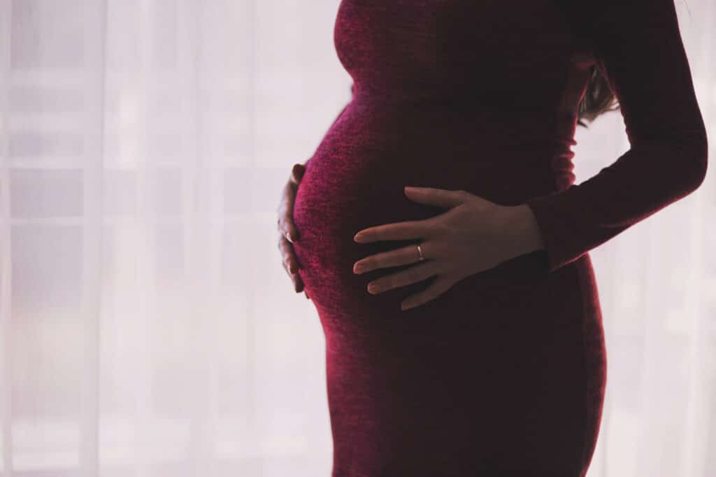 A pregnant woman in a fitted burgundy dress cradles her bump with both hands, standing in front of a sheer white curtain. Her engagement ring is visible on her left hand.