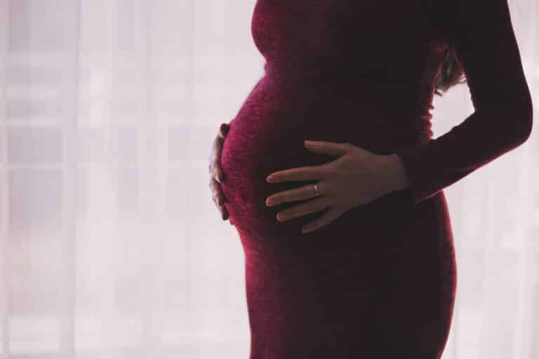 A pregnant woman in a fitted burgundy dress cradles her bump with both hands, standing in front of a sheer white curtain. Her engagement ring is visible on her left hand.