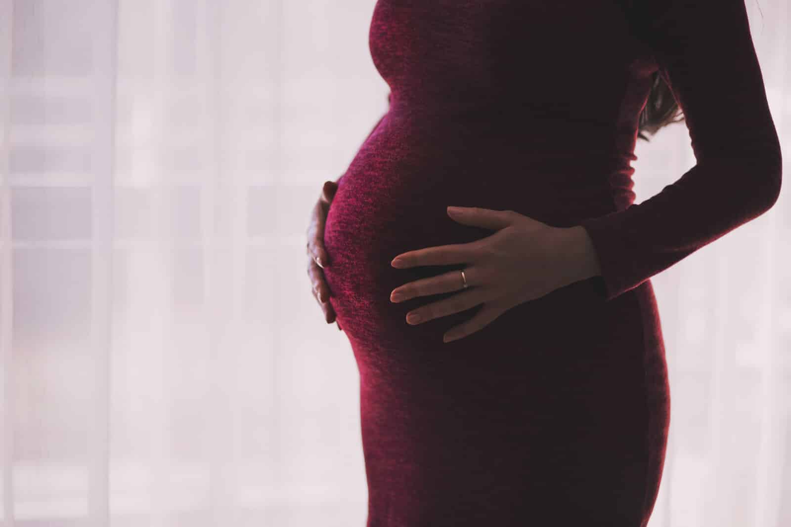 A pregnant woman in a fitted burgundy dress cradles her bump with both hands, standing in front of a sheer white curtain. Her engagement ring is visible on her left hand.