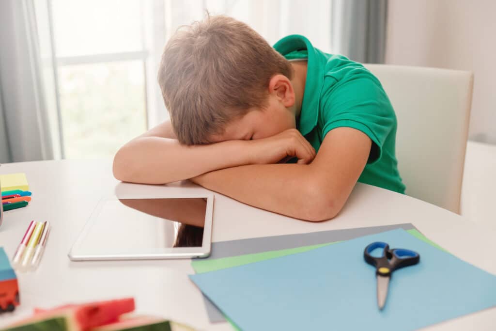 A young boy in a green shirt rests his head on his crossed arms at a desk. An electronic tablet, coloured pencils, scissors, and coloured paper are scattered on the table nearby.