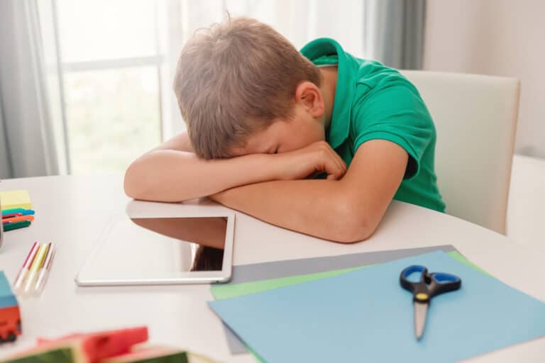 A young boy in a green shirt rests his head on his crossed arms at a desk. An electronic tablet, coloured pencils, scissors, and coloured paper are scattered on the table nearby.