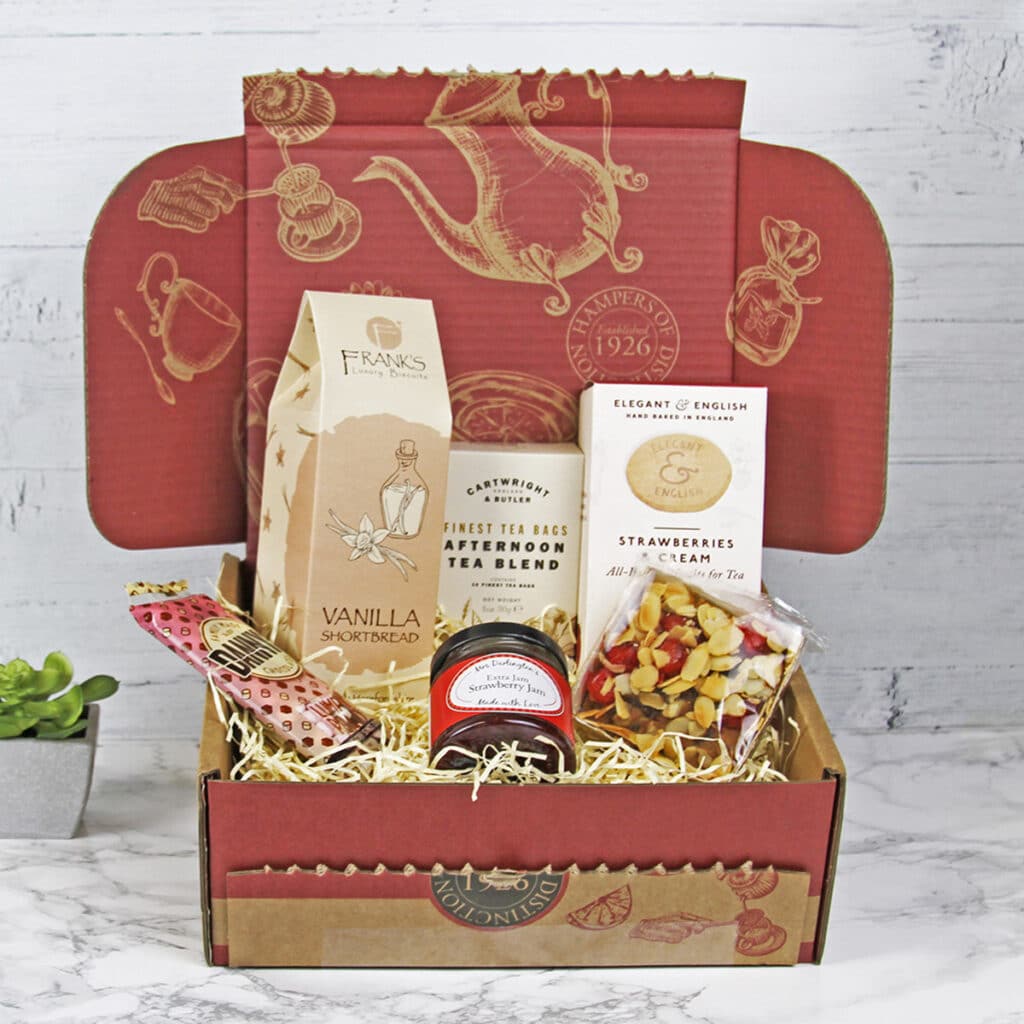 An afternoon tea hamper featuring tea, vanilla shortbread, strawberry biscuits, fresh fruit, and a jar of jam arranged on straw in a red decorative box, set on a marble surface with a small potted plant nearby.