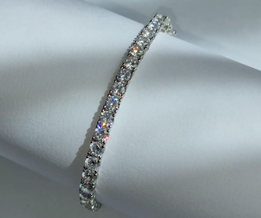 A sparkling tennis bracelet featuring a single row of round, clear diamonds set in silver, displayed on a rolled white surface with light reflecting off the stones—one of the essential accessories for brides on their wedding day.