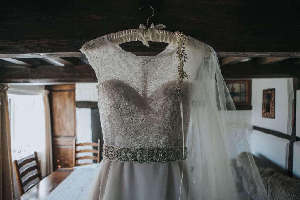 A white lace wedding dress with a jewelled belt and sheer veil hangs on a hanger in a rustic, wooden-beamed room with natural light coming through the windows.
