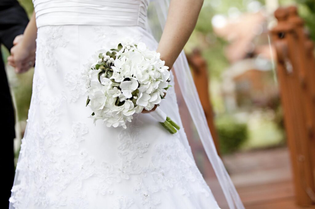 A bride in a white wedding dress holds a bouquet of white flowers outdoors, holding hands with someone special—a perfect moment that highlights the types of professional assistance you need when planning your wedding. A wooden structure is blurred in the background.