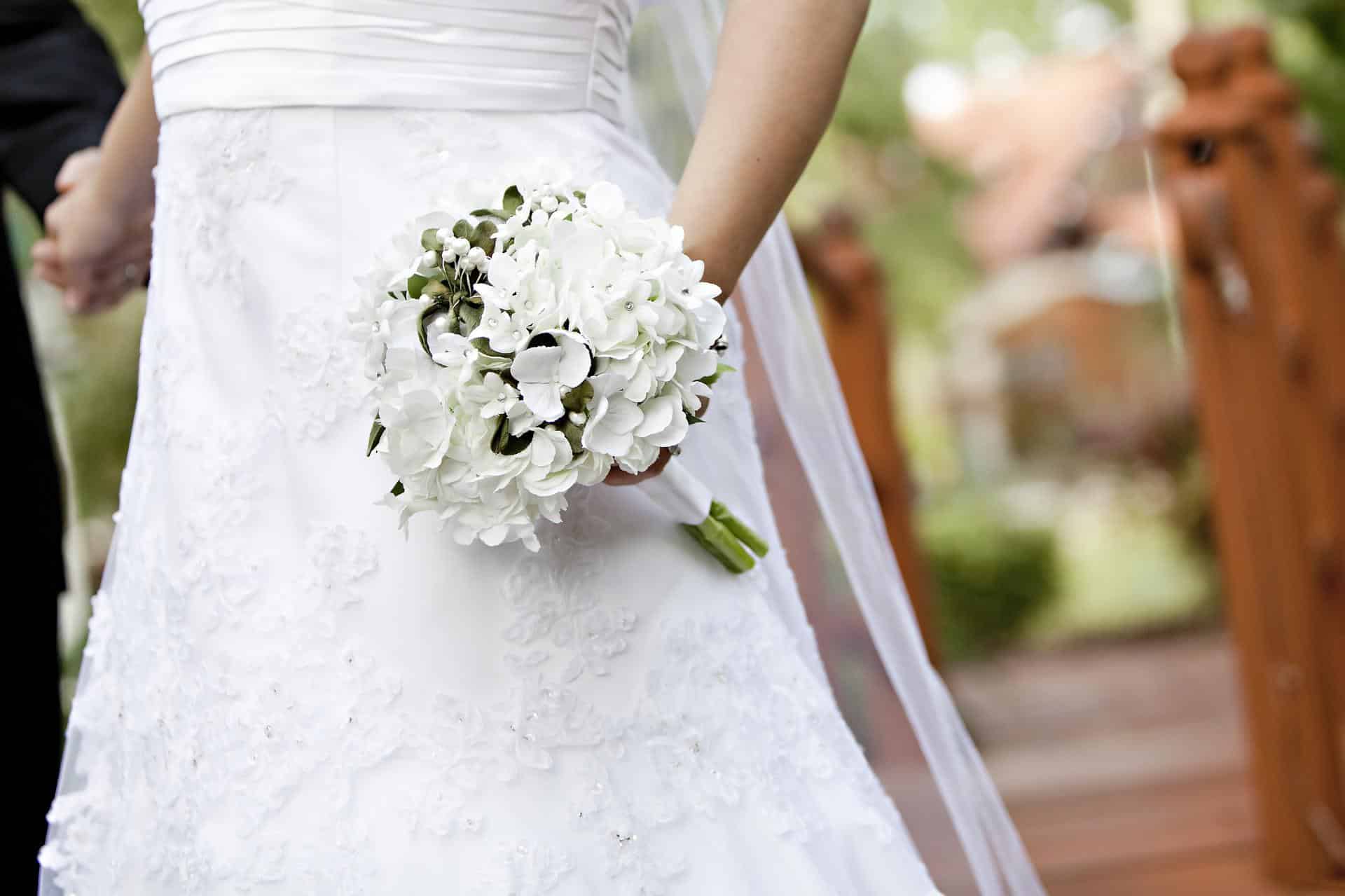 A bride in a white wedding dress holds a bouquet of white flowers outdoors, holding hands with someone special—a perfect moment that highlights the types of professional assistance you need when planning your wedding. A wooden structure is blurred in the background.