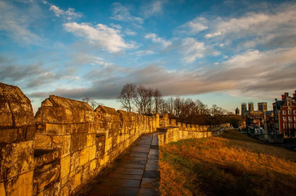A stone walkway runs along the York Bar walls at sunset, with golden light illuminating the stones and grassy embankment—a perfect scene for A Guide to the Ultimate Family Day Out in York, with distant trees and historic buildings under a partly cloudy sky.