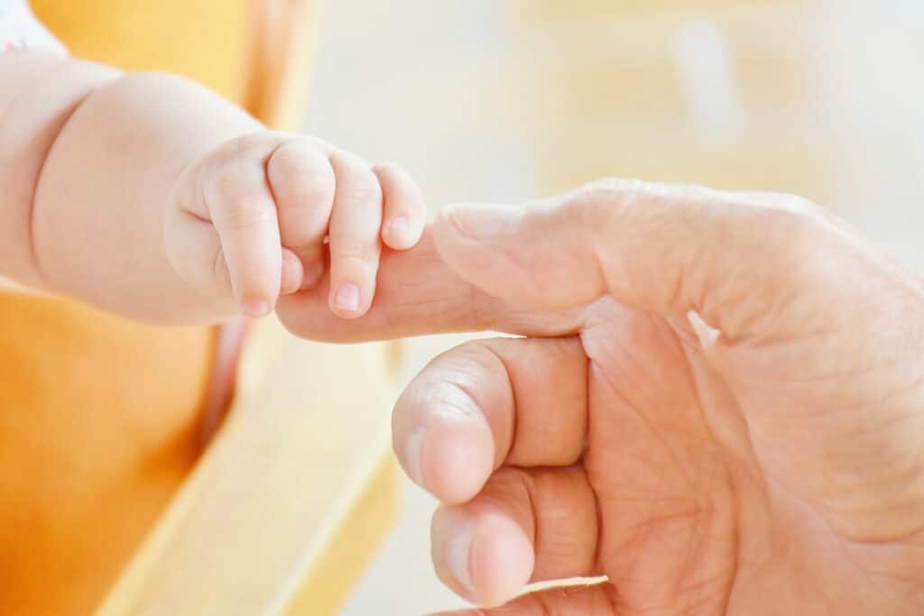 A baby's hand gently grasps an adult's finger against a soft, blurred background, symbolising care, connection, and tenderness.