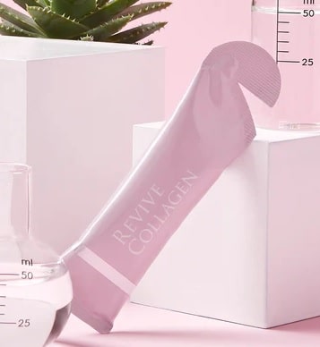 A pink sachet labelled "Revive Collagen" rests against a white cube, with lush greenery and glass beakers filled with clear liquid in the background, all set on a soft pink surface to highlight the benefits of revive collagen.