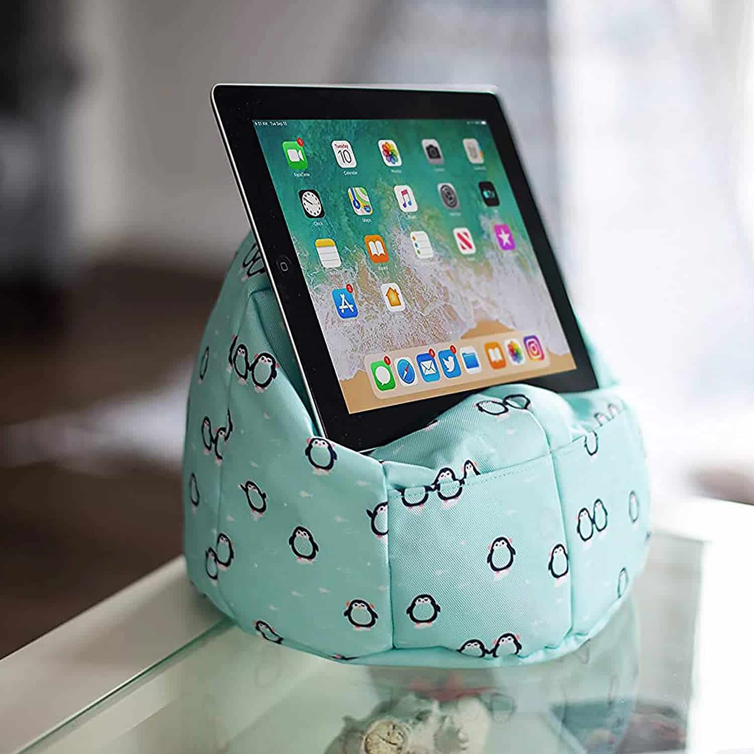 A tablet rests on a light blue, bean bag-style Planet Buddies stand with a pattern of small black circles, placed on a glass table indoors.