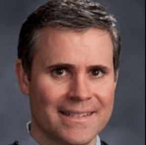 A man with short brown hair and grey eyes smiles slightly whilst wearing a dark suit jacket, light shirt, and tie. The background is a soft, blurred grey—like the calm before The Carnival: A Fun Way To Experience St John.