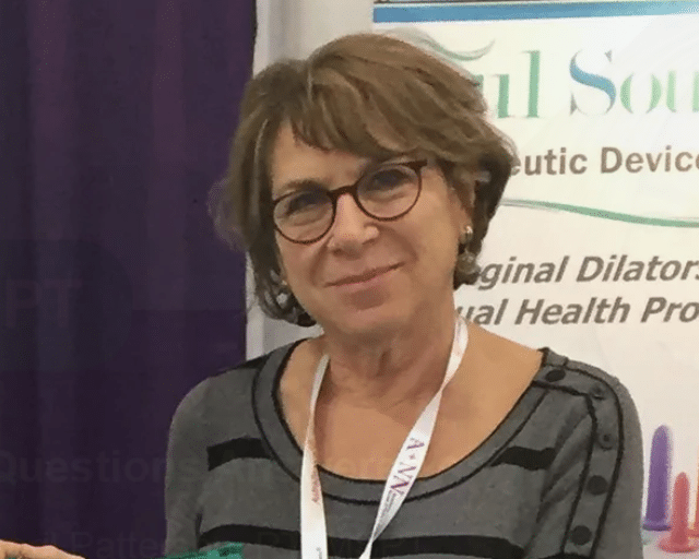 A woman with short brown hair and glasses, wearing a striped top and a white lanyard, stands in front of a stand display featuring the question, "Do I really need to be stretching?" with partially visible text.